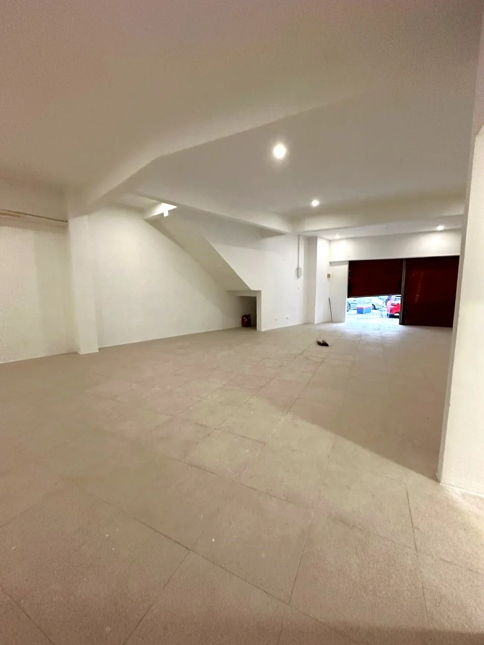 Room features: white painted walls, grey tiled flooring, high ceiling, recessed downlights, large open space, staircase leading to upper level, two large roll-up doors, no visible furniture or decor; style: modern.